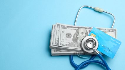 A hero image for an article titled "The Wealth-Health Connection: How Economic Status Shapes Well-Being". It is an image with a light blue background and pictures a stethoscope wrapped around a pile of US dollars and a credit card, signifying wealth and health.