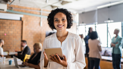 A photo of a woman standing in an office environment, smiling, and holding a tablet. She has tan skin and short dark curly hair, and is wearing a white long-sleeved shirt.