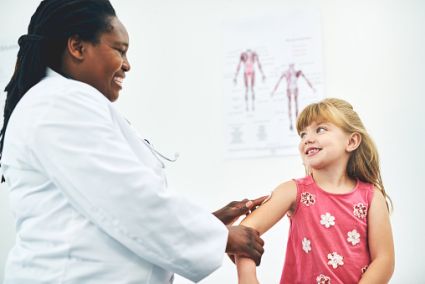 An image of a woman doctor with brown skin and long black hair, wearing a white lab coat, tending to a girl child patient with white skin, blonde hair with bangs, and wearing a pink shirt with flowers on it.
