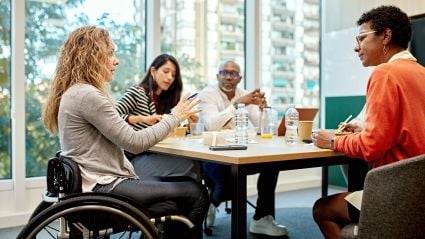 An image of four diverse adults sitting around a table having a discussion in an office space. One appears to be a woman with blonde curly hair, white skin, and a gray sweater sitting in a wheelchair. Another appears to be a woman with brown skin, short brown hair, glasses, and an orange shirt. Another appears to be a woman who is Asian, with white skin and brown hair wearing a striped top. And the other person appears to be a man with brown skin, a bald head, who is wearing business attire.
