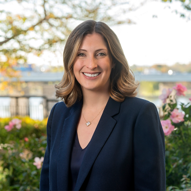 A smiling Caucasian female-presenting person with brown hair wearing a dark blue suit jacket against a green foliage background.