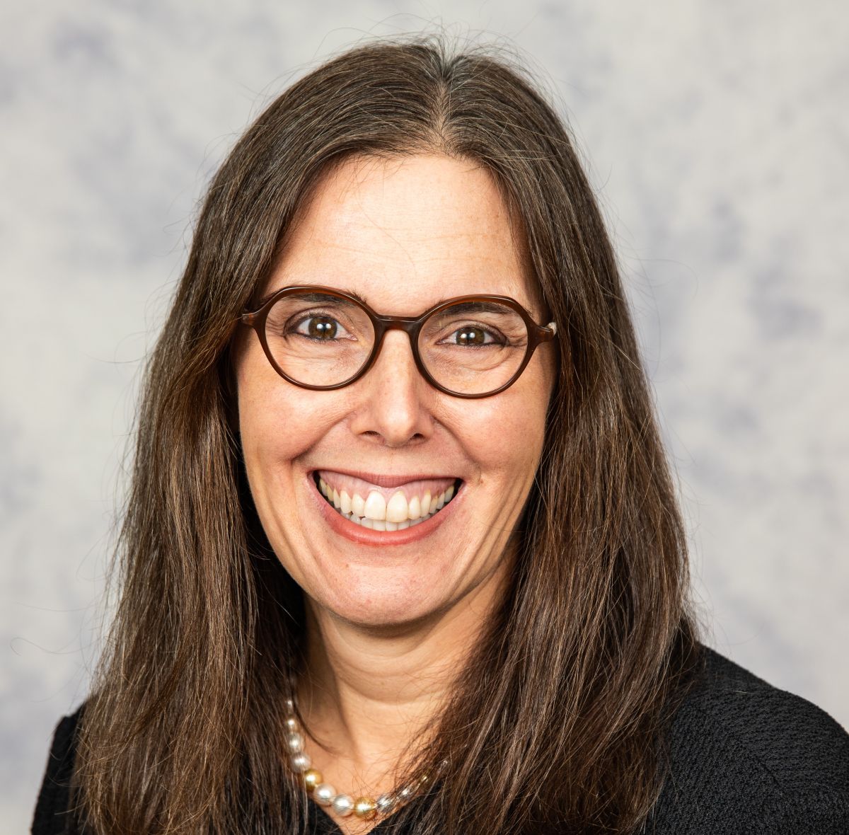 A smiling light-skinned female-presenting person with brown hair wearing a dark shirt against a grey background.