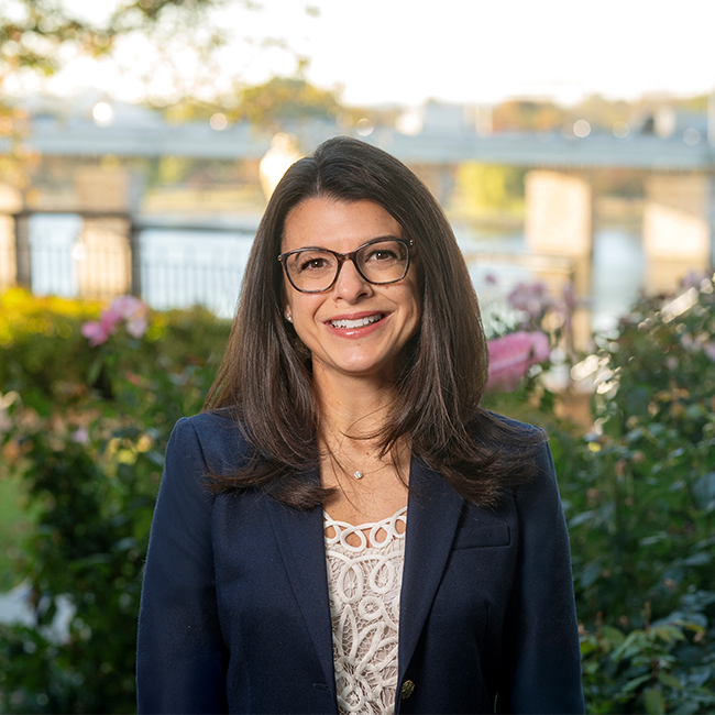Smiling light-skinned, female-presenting individual with long dark hair and glasses wearing a blue blazer against a nature-like background.