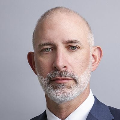 A headshot of a man named Andrew Milgram. He has a short white beard, white skin, and a bald head. He is wearing a suit and looking at the camera seriously against a gray background.
