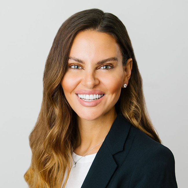 An image of a woman named Ashley Tyrner-Dolce, who is smiling at the camera against a white background. She is wearing a dark blazer with a white shirt and stud earrings, and she has white tanned skin and long brunette hair with hazel eyes.