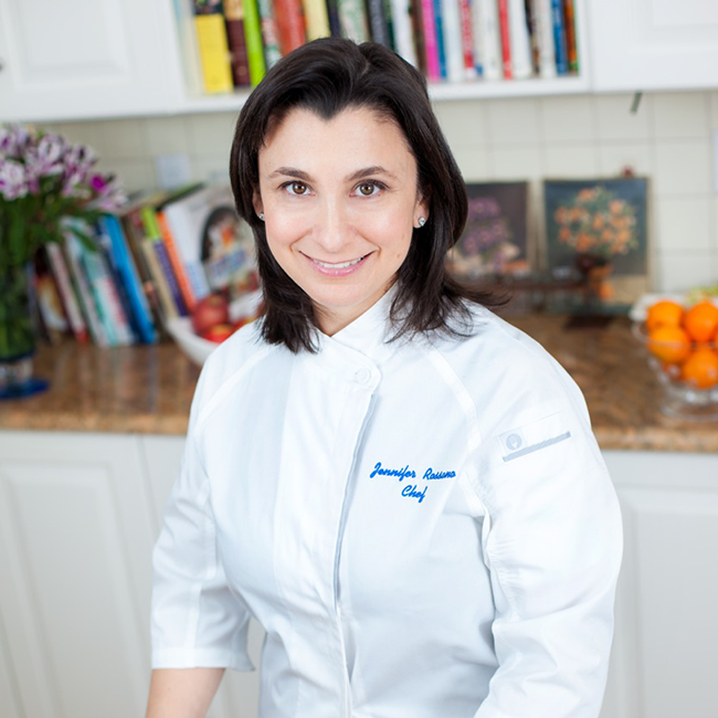 An image of a woman named Jennifer Rossano. She is smiling at the camera and standing in a kitchen in front of a shelf and counter of various cookbooks and miscellaneous items. She has light white skin, and shoulder-length dark brown hair and brown eyes. She is wearing a white outfit that appears to be a chef's uniform with her name printed in blue on the breast pocket. 