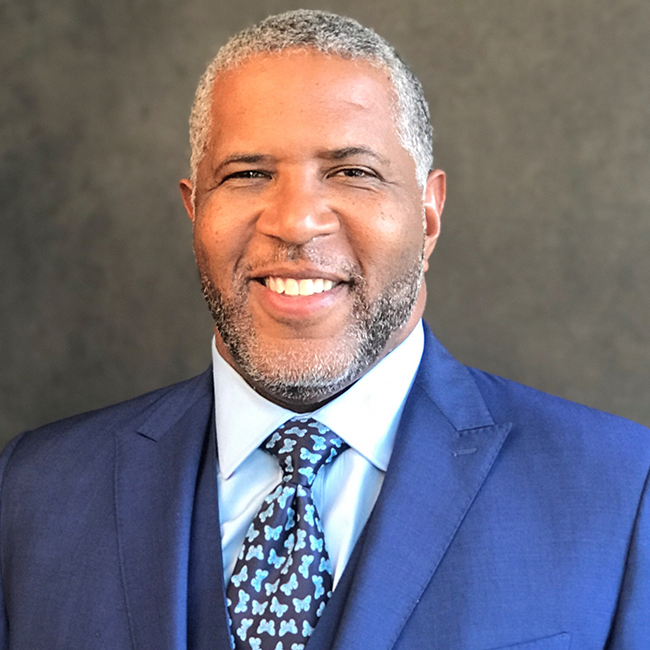 An image of a man named Robert F. Smith, who is smiling at the camera against a gray background. He has light brown skin, and grey-ish black short hair along with a beard. He is wearing a blue suit with a collared shirt and a patterned blue tie.