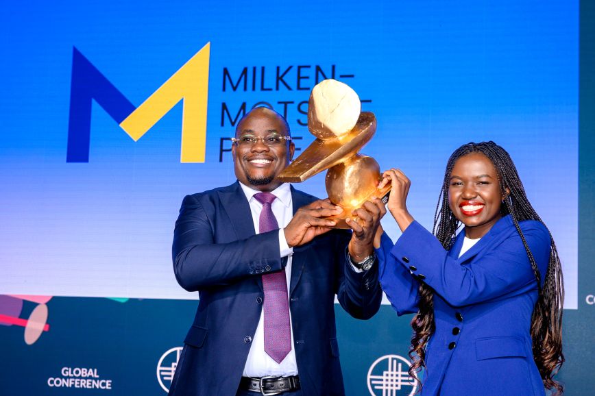A Black man and Black woman receive an award for winning a competition while standing on a stage at a big event