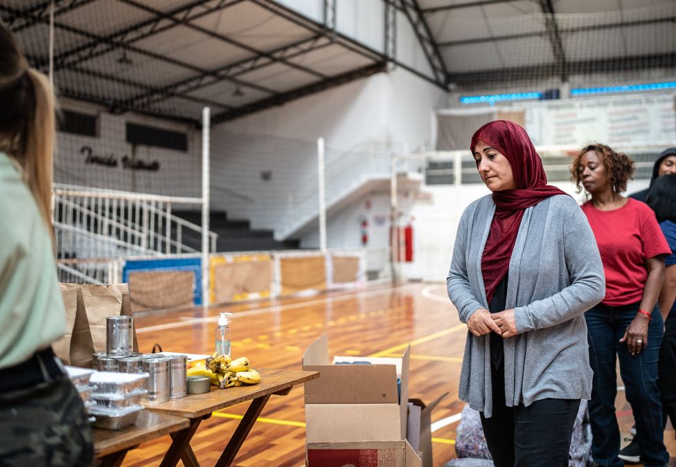 An image of various adults standing in a line in what appears to be a school gymnasium and waiting to collect food from a table, indicating a food bank or food drive.